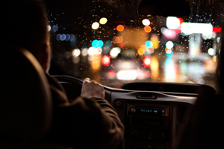 En person sitter bakom en ratt och kör bil i mörkret, genom vindrutan syns olika nattljus i oskärpa