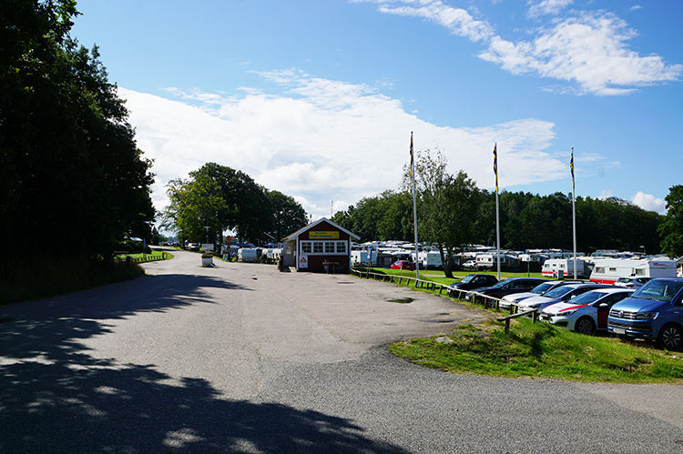 entrén till Unda camping en somrig dag, det är blå himmel och man ser parkerade bilar, ett litet rött receptionshus och husvagnar i bakgrunden 