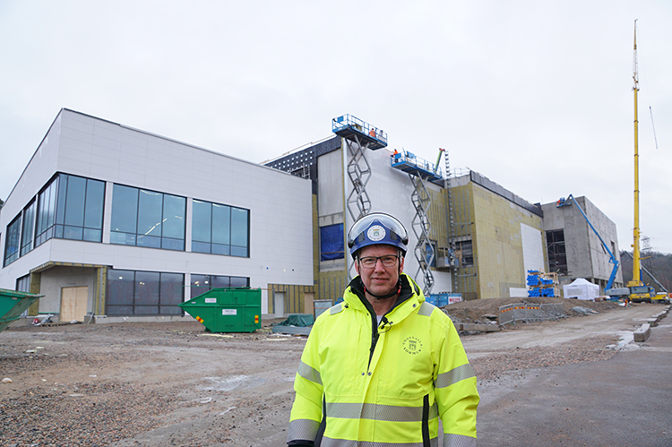 Anders Emilsson står framför en stor byggnad med vit fasad, och ett par kranar i bakgrunden 