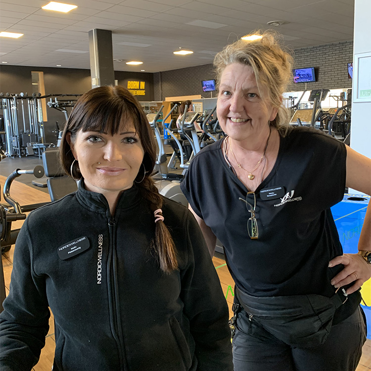 Två kvinnor står framför en gymlokal med träningsredskap