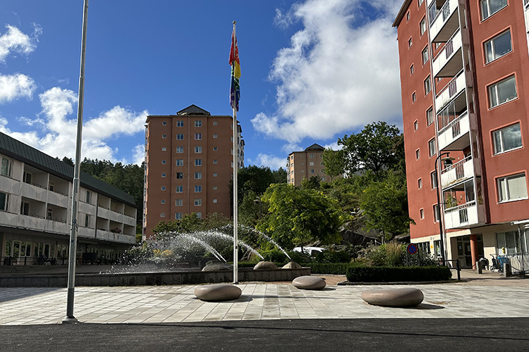 Ett torg med flata stenar, en fontän och ett par högre hus vid sidan i mitten syns en flaggstång med olika färger på en flagga