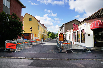 En asfalterad gata med byggstaket runtom på sidan, man ser ett gult hus och ett vitt hus med röd rundad markis