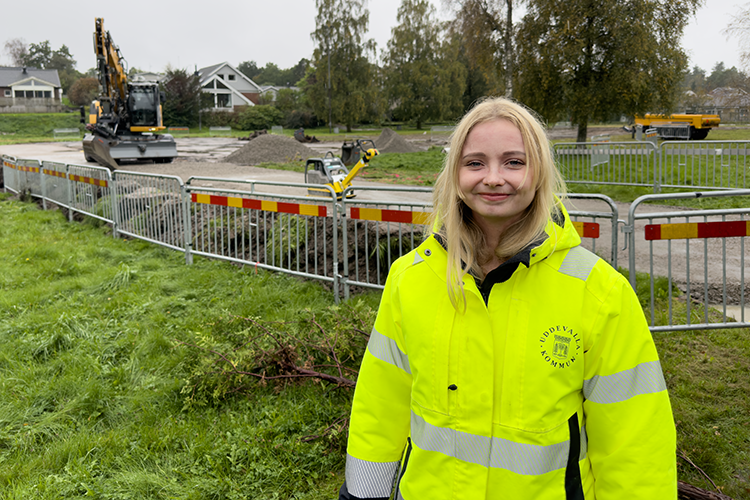 Madelene Petersson står i gula varselkläder framför ett arbetsområde med grävmaskin