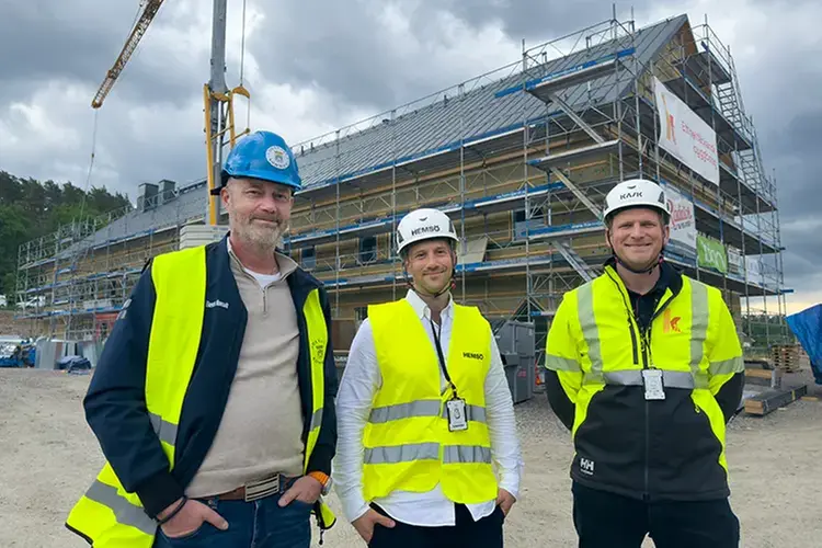 tre glada män i bygghjälmar och varselvästar står framför ett hus med byggnadsställningar