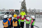 En man i varselkläder och bygghjälm pratar med många yngre barn som har bygghjälmar på huvudet och vinterkläder, det är snö ute
