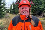 En kvinna står i orange arbetskläder och hjälm med visir i ett skogsområde